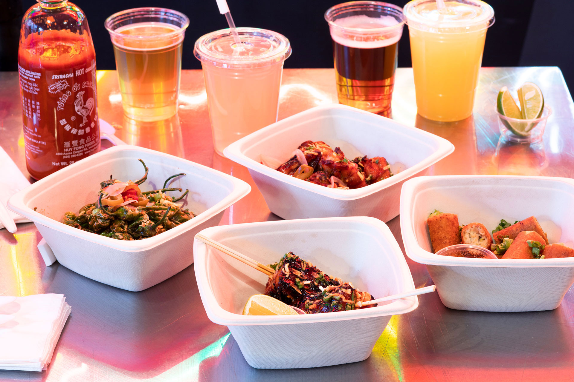 an assortment of appetizers from sari sari including lumpia, shishito peppers, and skewers, set against multiple beers and fresh juices